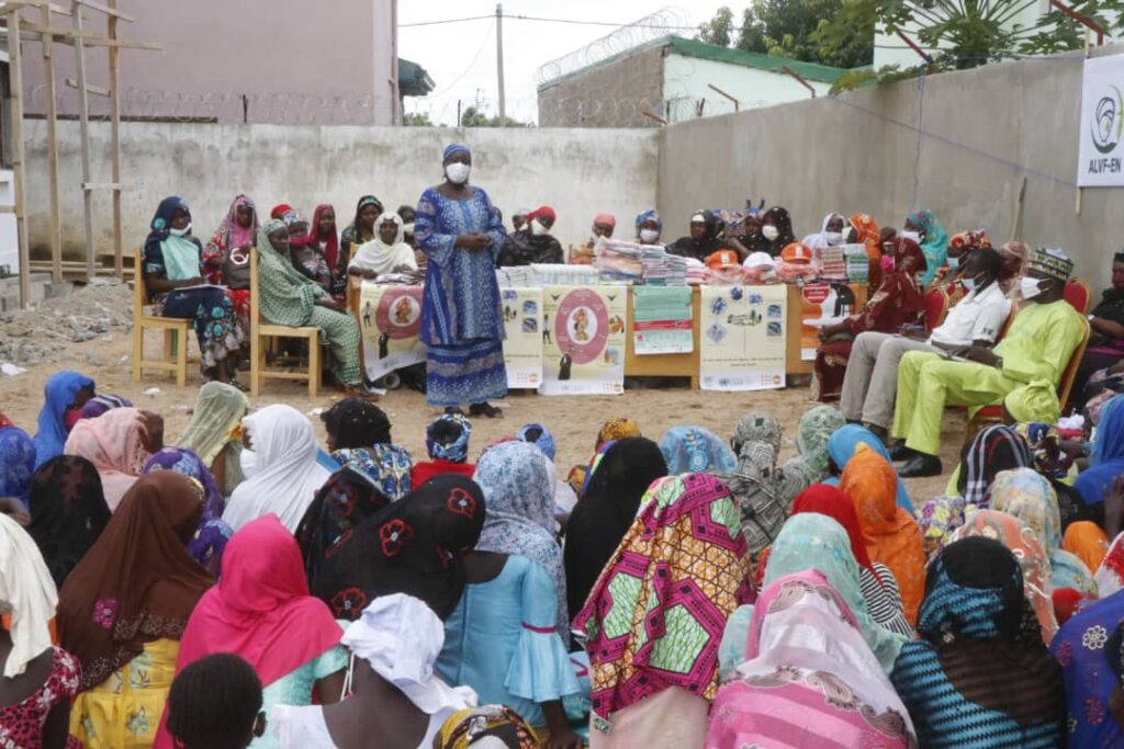 A group of girls and young women, many wearing colorful headscarves, are gathered outdoors for an educational session. A woman in a blue traditional outfit and mask, Aissa Doumara, leads the discussion, standing in front of posters and tables filled with educational materials.