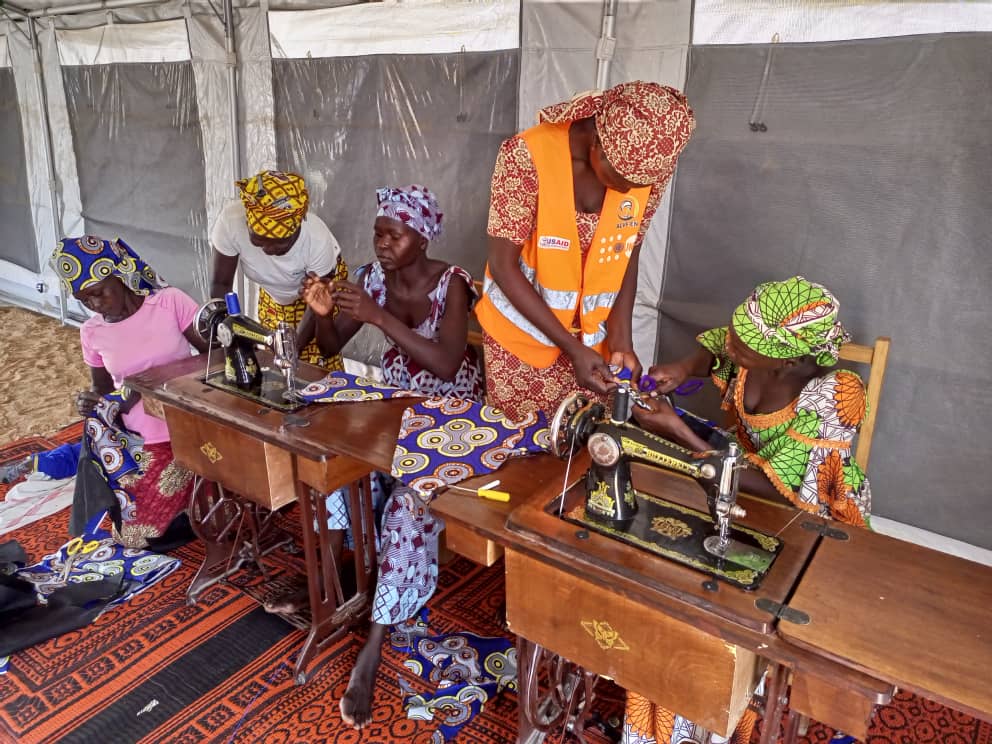 Quatre femmes survivantes de violences basées sur le genre, ainsi qu'une facilitatrice portant un gilet orange, sont assises devant des machines à coudre à l'intérieur d'une tente. Elles participent à un atelier de couture visant à développer des compétences et à les autonomiser, en utilisant des tissus aux motifs colorés.