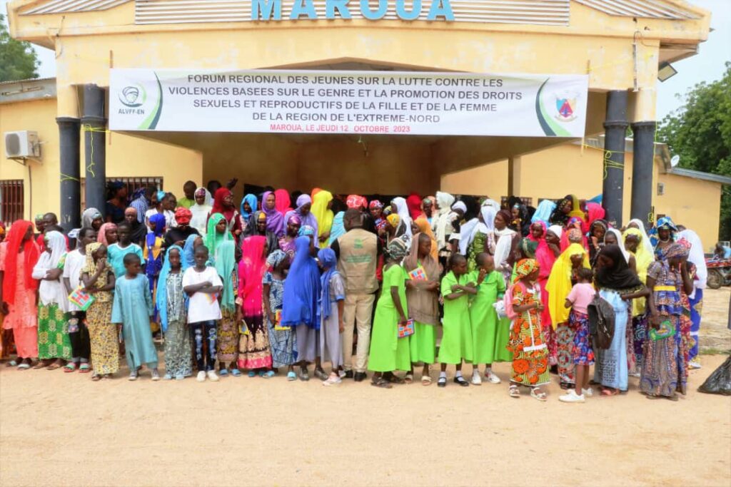 A large group of activists, mostly young girls and adolescents, stand together in front of a building with a sign displaying the name of the forum: 'Forum Regional des Jeunes Sur la Lutte Contre Les Violences Basees sur le Genre et la Promotion des Droits Sexuels et Reproductifs de la Fille et de la Femme de la Region de L'extreme-nord". The participants are gathered to raise awareness about sexual and gender-based violence.