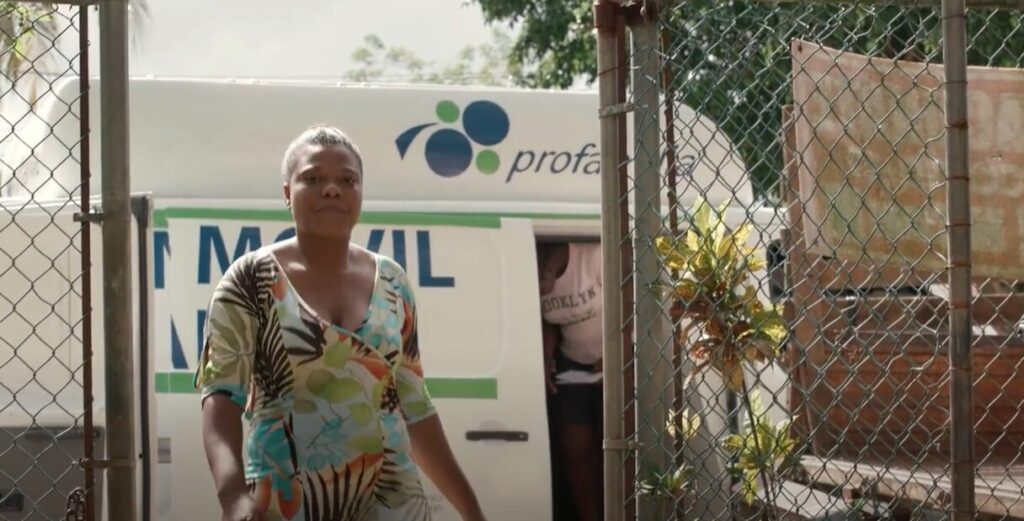 A woman stands in front of a Profamilia mobile health unit, which is parked behind a chain-link fence. She is wearing a brightly patterned dress and looks forward confidently as she walks. Another person is partially visible exiting the mobile unit behind her. The scene takes place in a suburban area, with plants and greenery in the background.