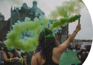 A person wearing a green bandana raises a green smoke flare in a crowd, creating a vivid cloud of green smoke in the air. The person’s raised arm and bandana symbolize solidarity and activism for reproductive rights The scene captures a powerful moment of collective action and resistance.