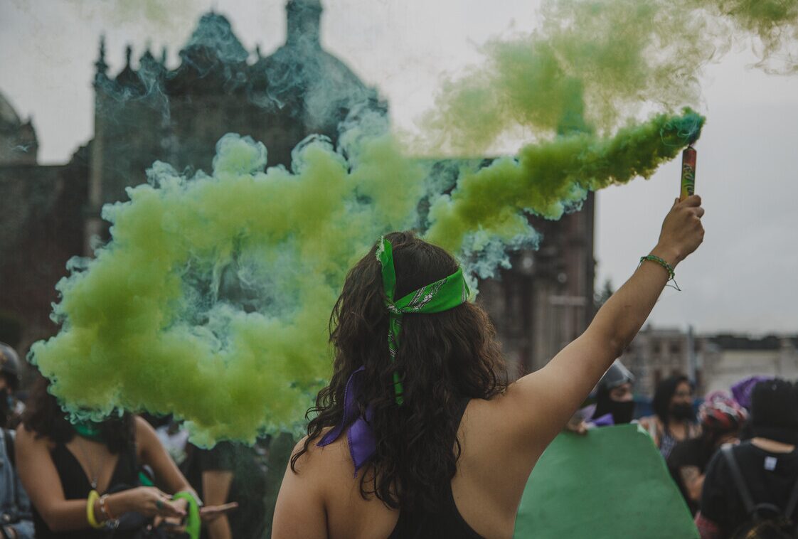 A person wearing a green bandana raises a green smoke flare in a crowd, creating a vivid cloud of green smoke in the air. The person’s raised arm and bandana symbolize solidarity and activism for reproductive rights The scene captures a powerful moment of collective action and resistance.